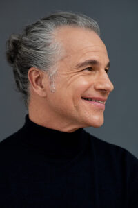 Bruno Pelletier souriant, posé de profil les cheveux attachés et portant un col roulé noir, sur fond gris. 