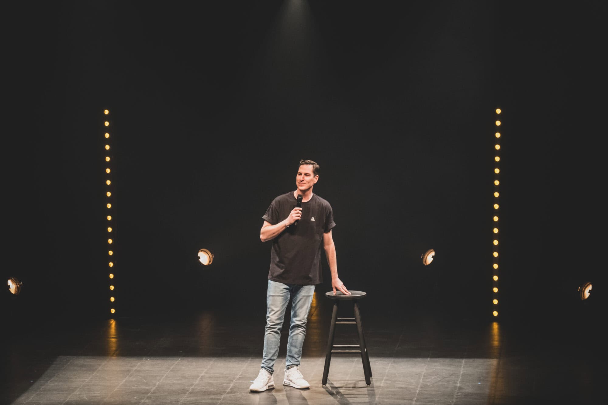 Guillaume Pineault en spectacle sur scène en formule stand up, micro en main et une main posée sur un tabouret